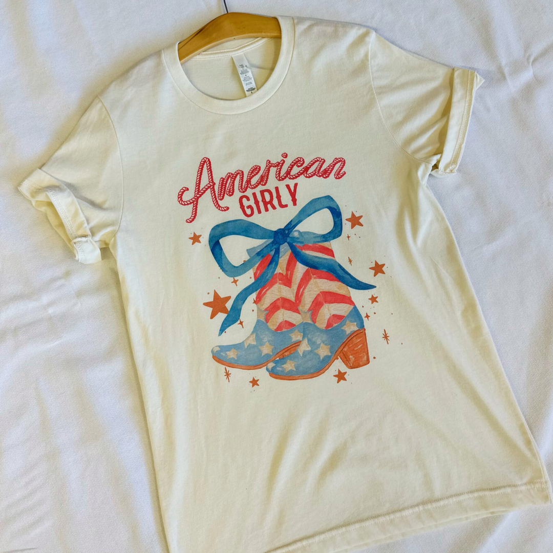 4th of july t shirt, american girly, pair of cowboy boots with stars and stripes like the american flag with a big blue bow on top and stars floating around the boots