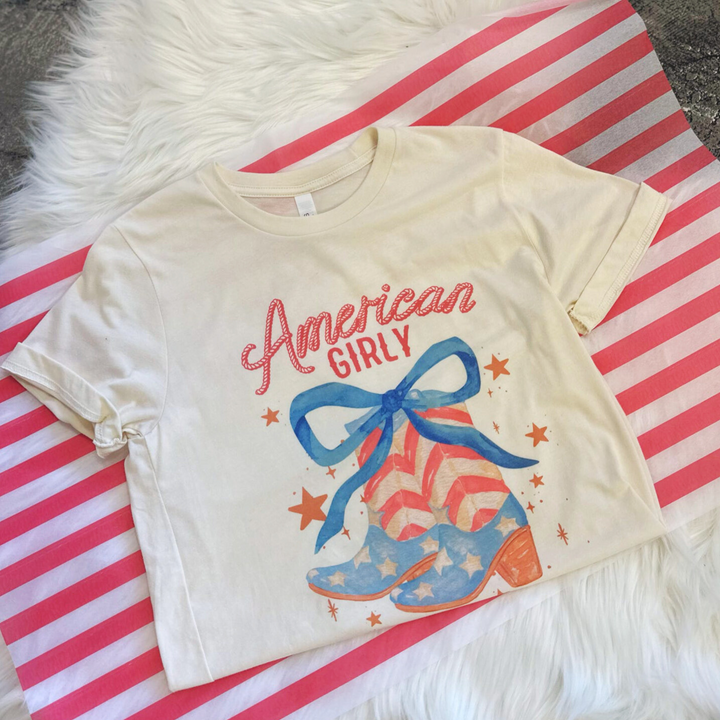 4th of july t shirt, american girly, pair of cowboy boots with stars and stripes like the american flag with a big blue bow on top and stars floating around the boots