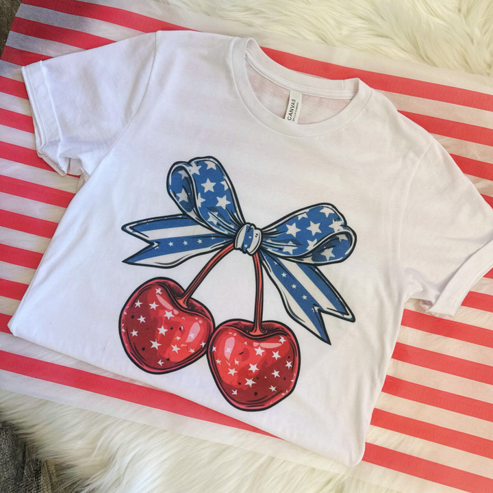 4th of july t shirt, white t shirt with a big pair of cherries on the front, red cherries with white stars, big blue bow on top of the cherries with navy and white stripes and stars