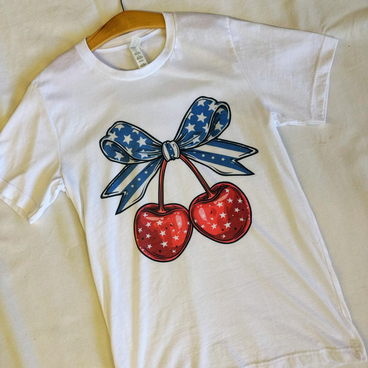 4th of july t shirt, white t shirt with a big pair of cherries on the front, red cherries with white stars, big blue bow on top of the cherries with navy and white stripes and stars