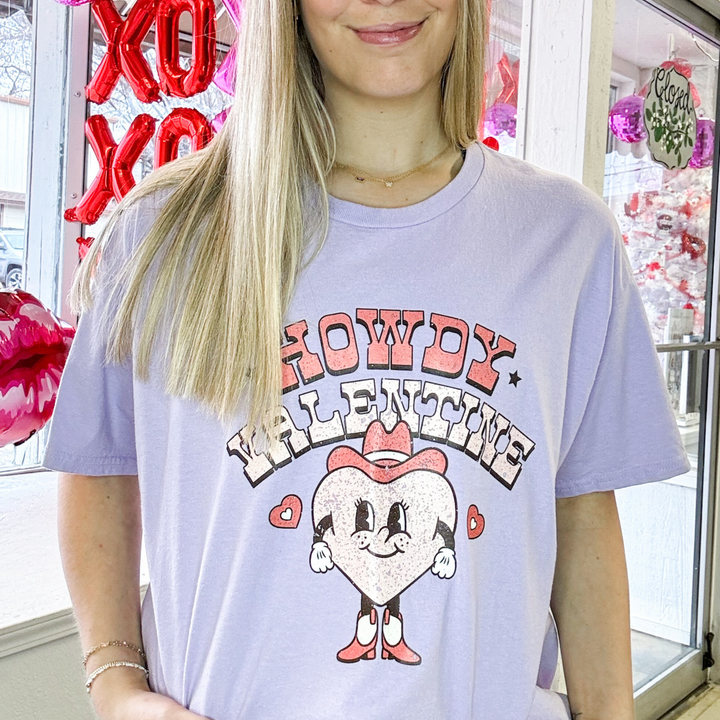 purple valentines graph t shrit that says howdy valentine in red and light pink font, beneath the writing is a heart with a cowboy hat and boots on