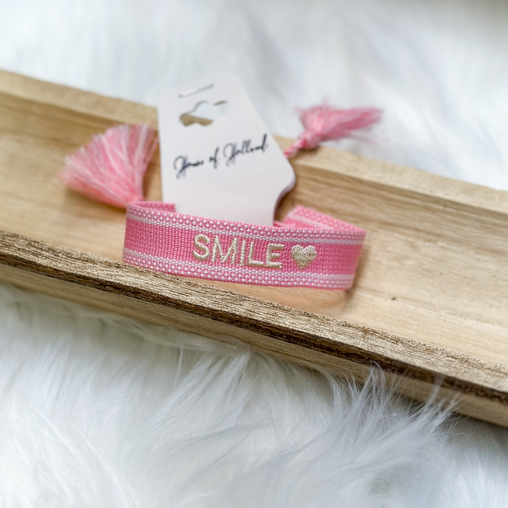 pink and white embroidered bracelet, the word smile with a heart at the end in white, adjustable with tassels at the end. 