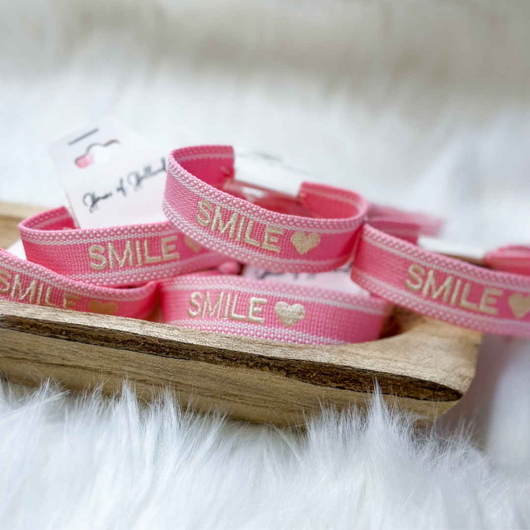 pink and white embroidered bracelet, the word smile with a heart at the end in white, adjustable with tassels at the end. 
