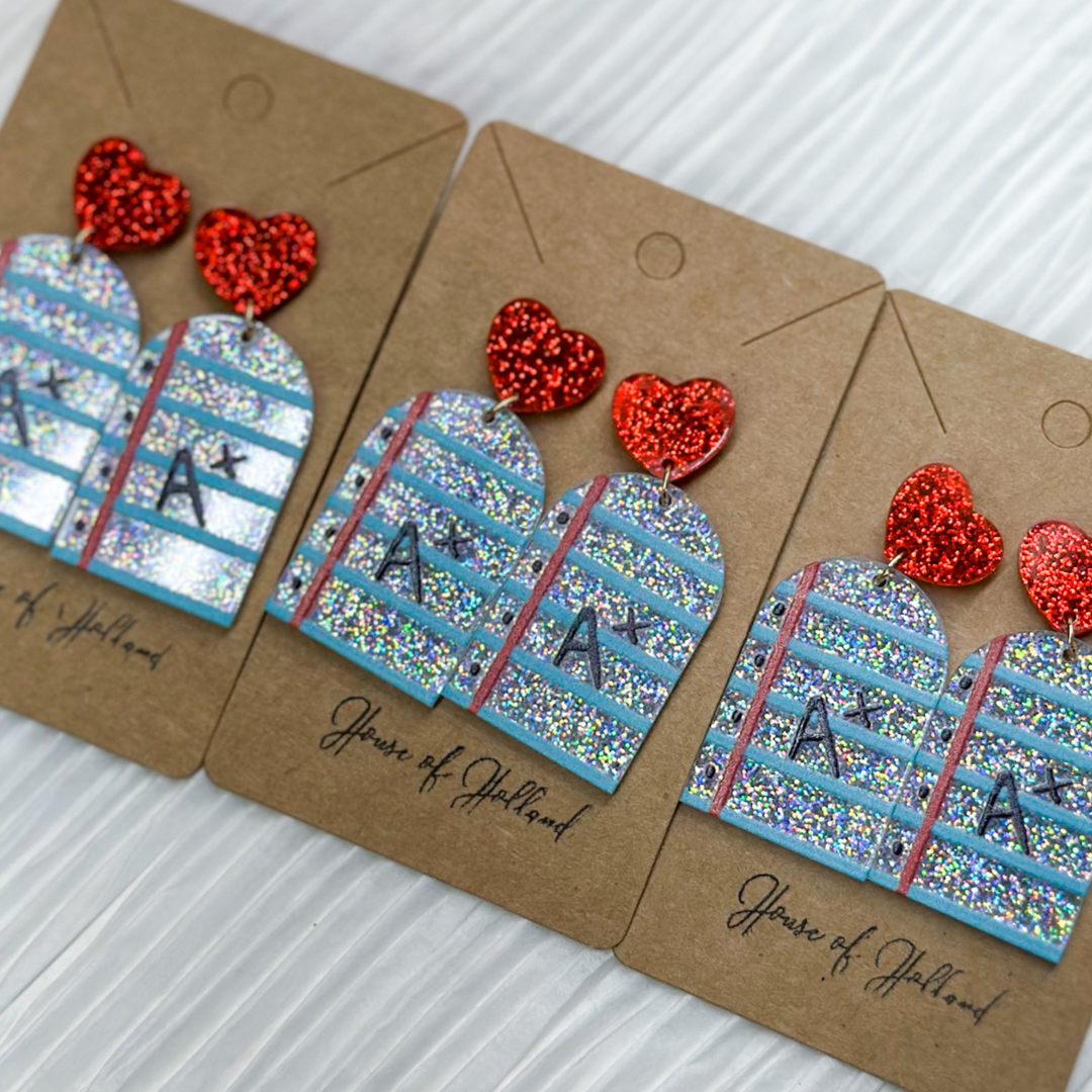 teacher earrings, acrylic, notebook paper with blue lines, silver glitter background, A+ in black dangling from a red glitter stud. 