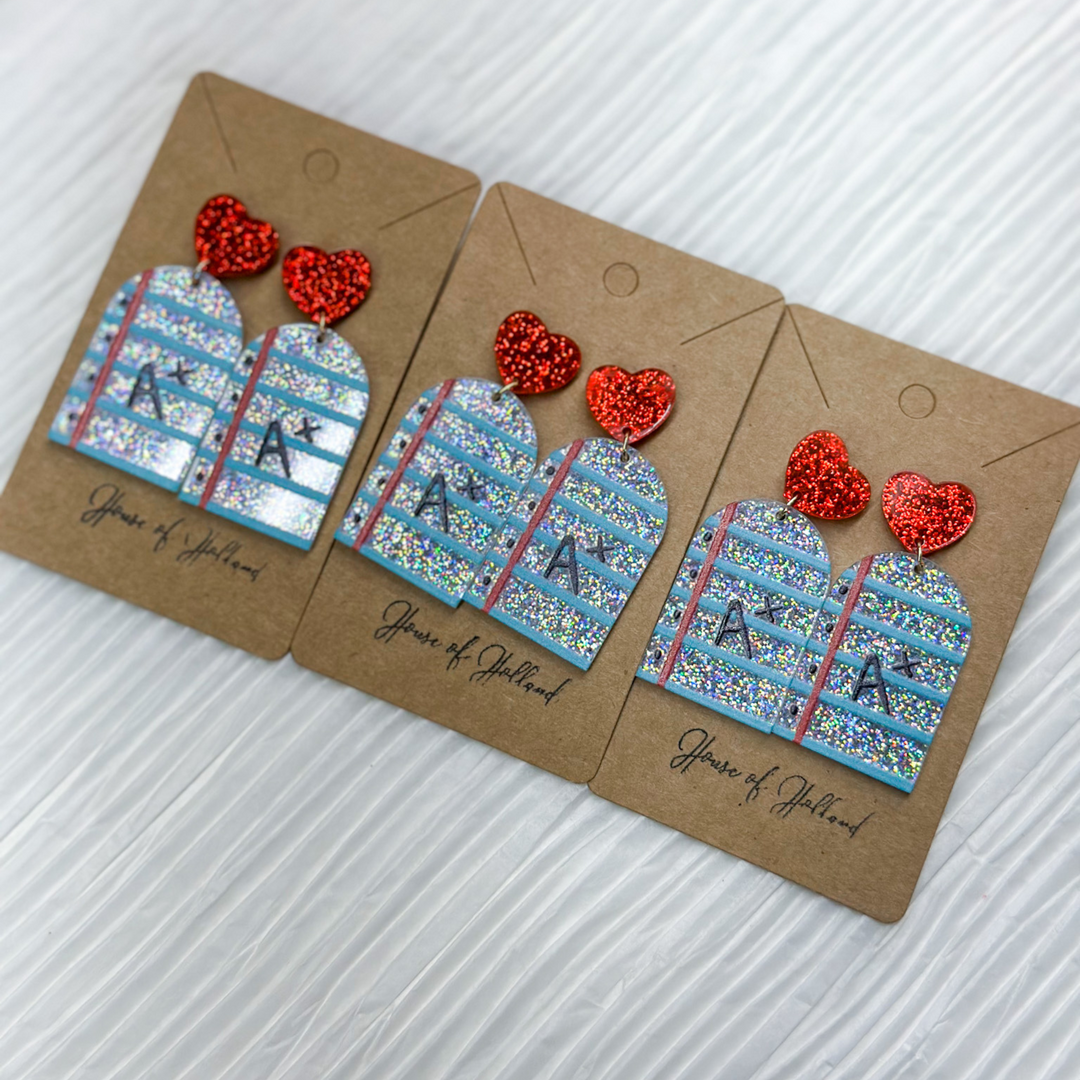 teacher earrings, acrylic, notebook paper with blue lines, silver glitter background, A+ in black dangling from a red glitter stud. 