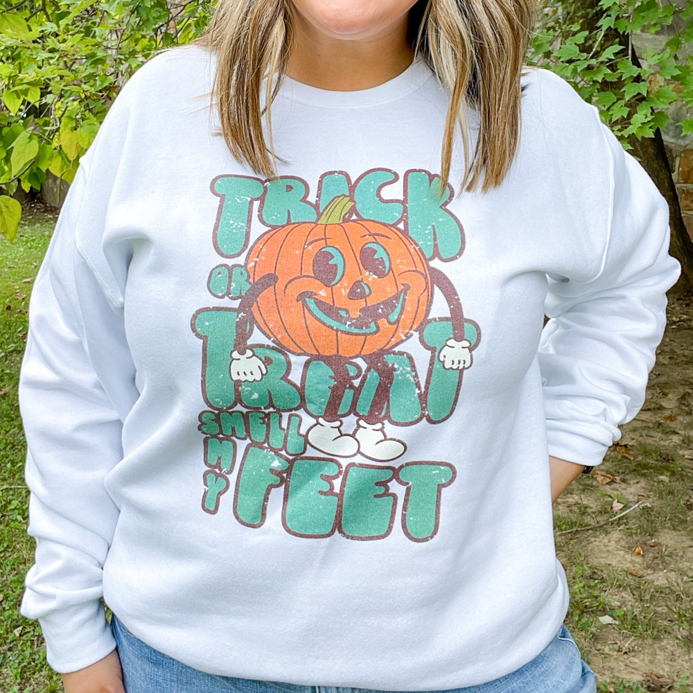 trick or treat smell my feet halloween graphic sweatshirt. white sweatshirt with green font and a big jack-o'-lantern sitting on the words. 