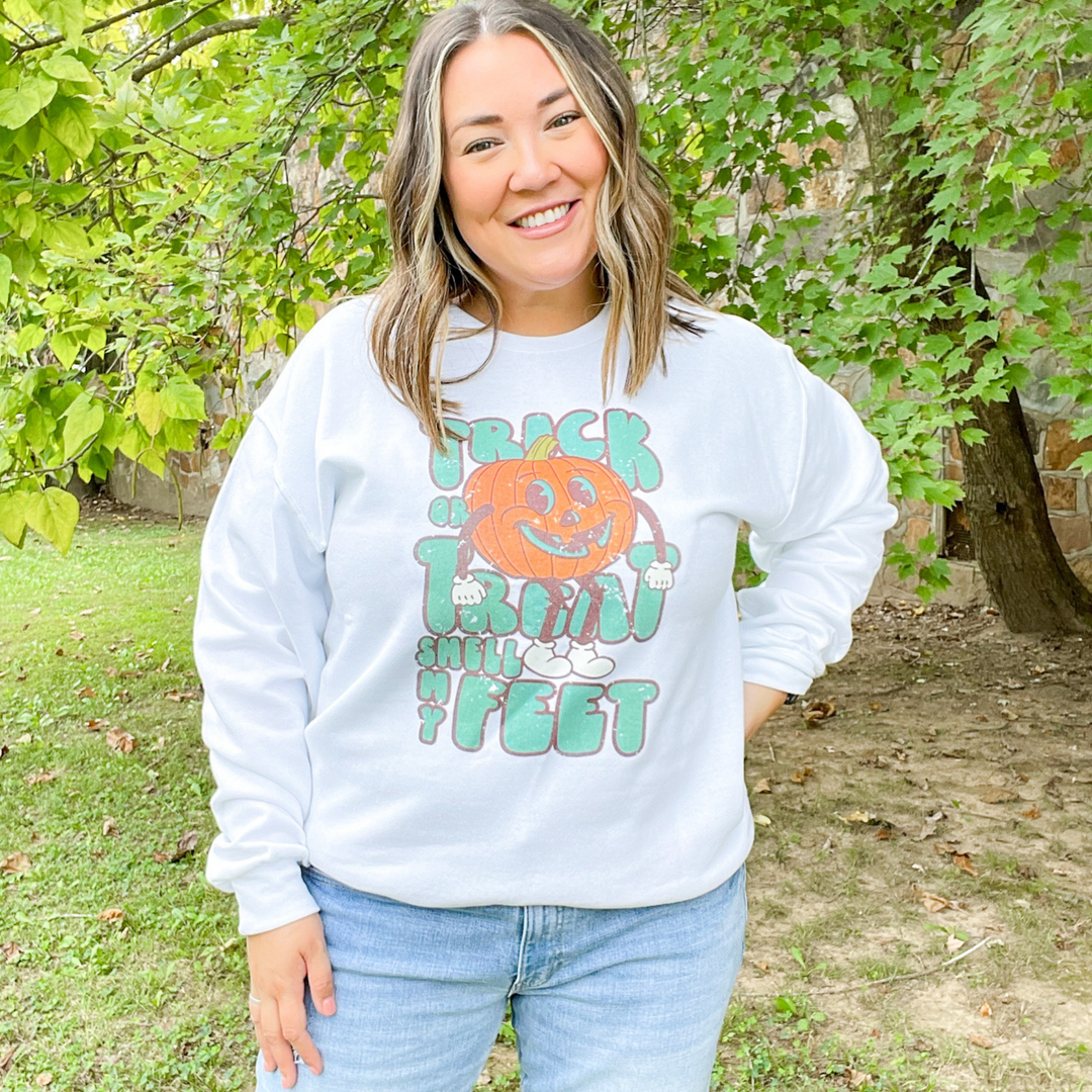 trick or treat smell my feet halloween graphic sweatshirt. white sweatshirt with green font and a big jack-o'-lantern sitting on the words. 